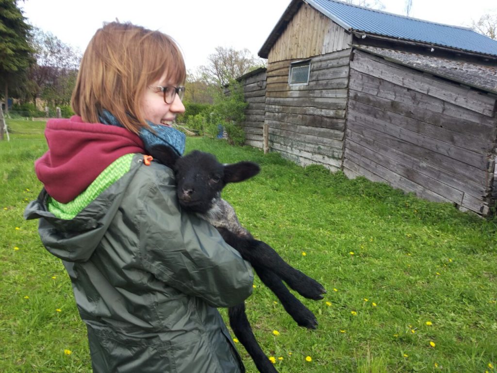 Auf einer grünen Wiese steht Astrid (vom fachwerkhuhn) und hält ein Lamm auf den Armen und strahlt dabei.