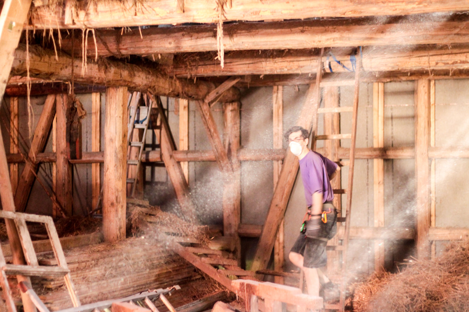 In der Scheune, die voll mit altem Holzund Heu ist, steht Lukas in Arbeitskleidung. Lichtstrahlen kommen durch das Dach.