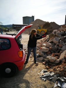 Katalin wirft auf einem Bauhof Steine aus dem Kofferraum des Autos auf einen großen Schutthaufen.